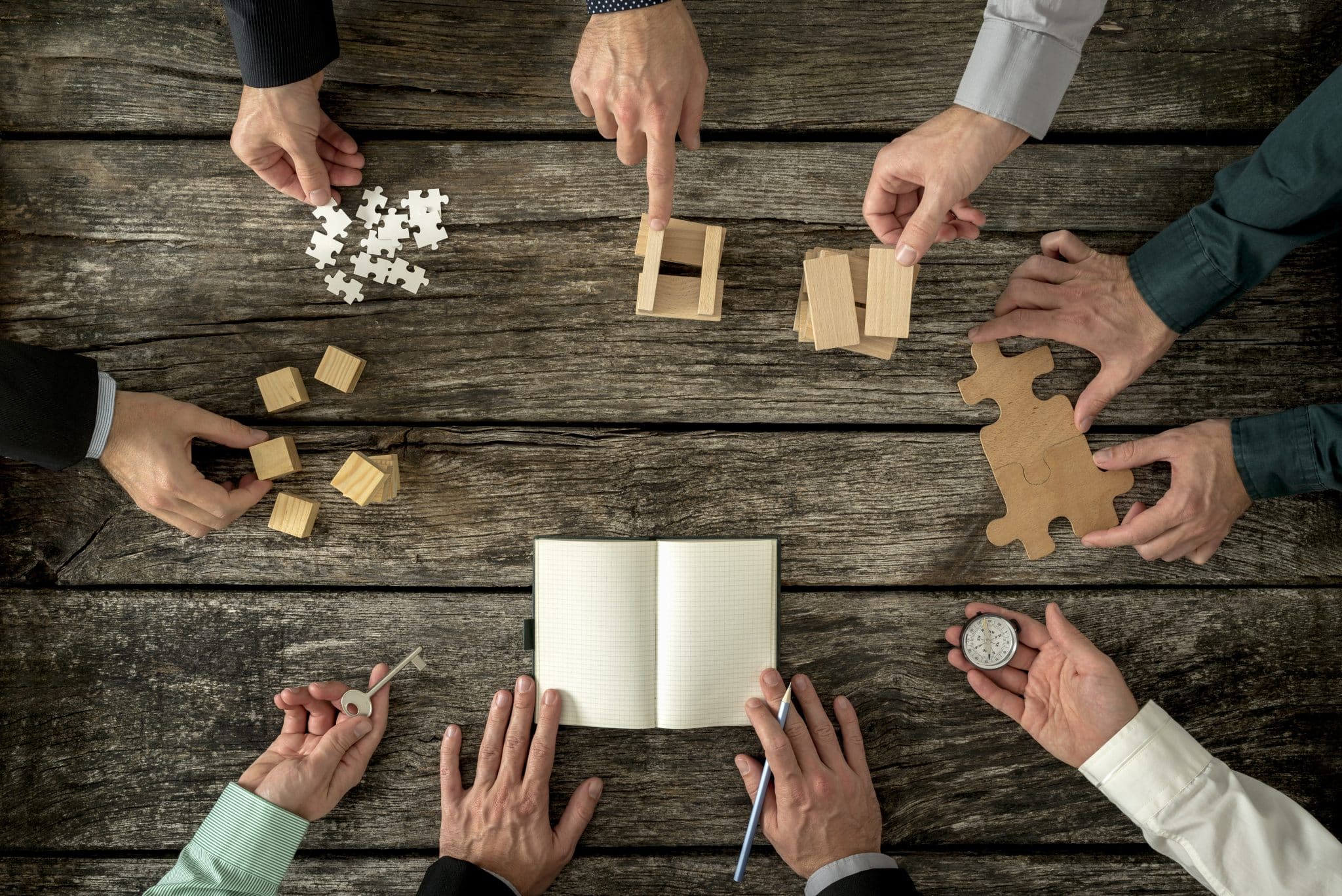 Eight businessmen planning a strategy in business advancement each holding different but equally important metaphorical element - compass, puzzle pieces, pegs, cubes, key and one making notes.