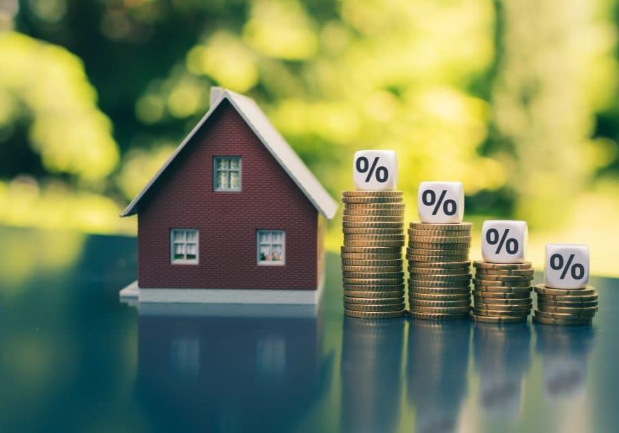 A small model home sits on a table next to stacks of coins with percentage symbols on decreasing high stacks of coins